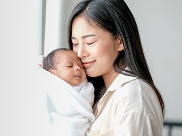 White shirt mother is kissing her newborn baby in bedroom in front of glass windows with white curtain to show love and family bonding.