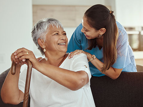 Happy, relax and senior woman with caregiver smile while sitting on a living room sofa in a nursing home. Support, help and professional nurse or healthcare worker helping elderly lady or patient.