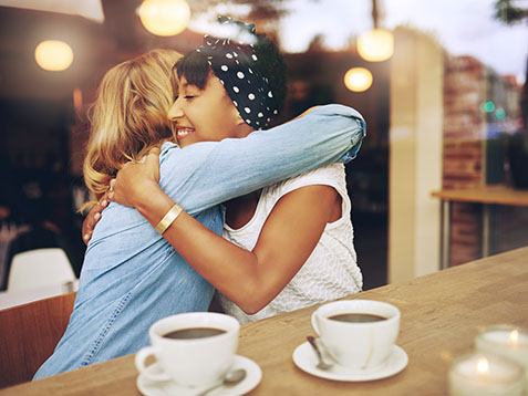 Two multi ethnic affectionate girl friends embracing as they sit in a coffee shop enjoying a cup of coffee together