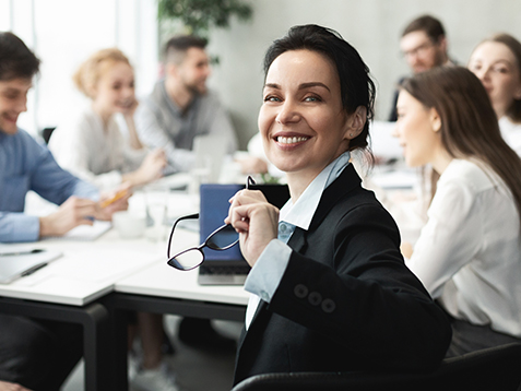 Top manager smiling to camera at meeting, colleagues discussing work on background, panorama