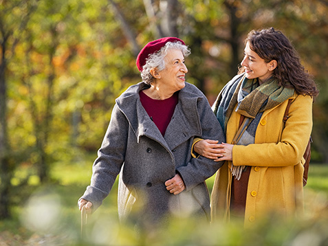 Young woman in park wearing winter clothing walking with old grandmother. Happy grandma wearing coat walking with lovely girl outdoor with copy space. Smiling lovely caregiver and senior lady walking in park during autumn and looking at each other.