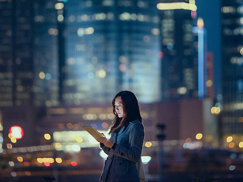 Pretty office lady is using digital tablet, standing against illuminated highrise corporate buildings at night time.