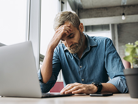 Stylish modern man with a beard in a blue shirt is sitting at the table and working in a laptop in the office