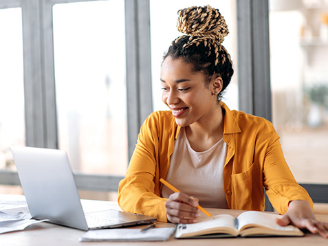 Online education, e-learning. Happy african american young woman in stylish casual clothes, studying remotely, using a laptop, listening to online lecture, taking notes while sitting at home, smiles