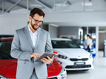 Smiling, friendly car seller standing in car salon and using tablet to check on new messages customers post on internet.