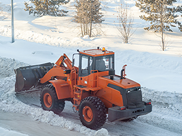 Big orange tractor cleans up snow from the road and loads it into the truck. Cleaning and cleaning of roads in the city from snow in winter