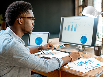 man wearing glasses looking at monitor screens with graphs on them