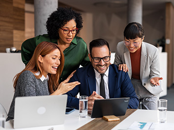 A group of professional colleagues collaborates in an office environment, sharing smiles and enthusiasm around a central laptop, depicting teamwork and connection in a corporate setting.