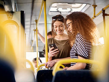 Two cheerful pretty young women are standing in a bus and looking at the phone and smiling while waiting for a bus to take them to their destination.