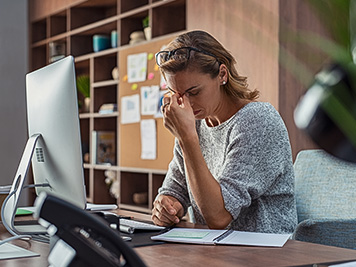 Exhausted businesswoman having a headache in modern office. Mature creative woman working at office desk with spectacles on head feeling tired. Stressed casual business woman feeling eye pain while overworking on desktop computer.