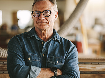 Portrait of successful senior man in eyeglasses standing in his carpentry workshop. Proud carpentry workshop owner standing with his arms crossed.