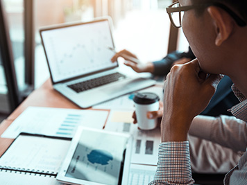 Business partnership pointing to the graph of the company financial statements report and profits earned during in the computer screen with giving a presentation to colleagues holding tablet and coffee in office room.