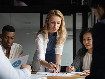 Concentrated young businesswoman explaining market research results in graphs to mixed race colleagues. Focused group of diverse employees holding brainstorming meeting, discussing project ideas.