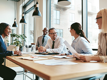 Diverse group of businesspeople laughing while discussing paperwork together during a meeting around a table in a modern office