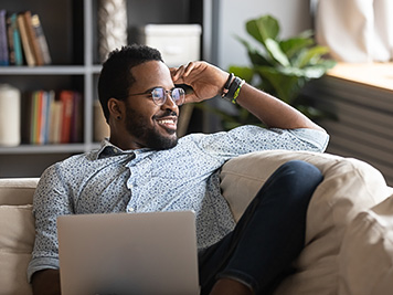 Happy young african american man stretching back, enjoying free leisure weekend time alone on sofa with computer. Smiling multiracial freelance guy distracted from job study, looking at window.