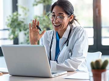 Shot of female doctor waving and talking with colleagues through a video call with a laptop in the consultation.