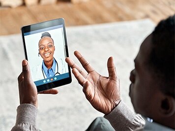 Middle aged African American man chatting with virtual female doctor at home on a tablet, telehealth consulting