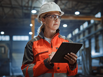 Professional Heavy Industry Engineer/Worker Wearing Safety Uniform and Hard Hat Uses Tablet Computer. Serious Successful Female Industrial Specialist Walking in a Metal Manufacture Warehouse.