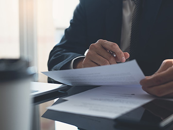 Signing contract, business agreement and deal concept. Closeup businessman in black suit reading before signing business contract documents at desk with laptop computer, busy with paperwork. Man manager proofing report at office