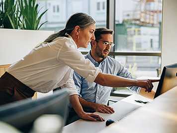 Mature woman working with young man sitting at desk and showing something on computer screen in office. Female executive pointing at desktop monitor and talking with male colleague in office.