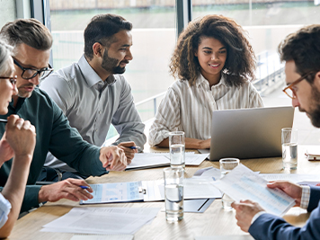 Diverse executive businesspeople discuss corporation financial plan at boardroom meeting table. Multiracial team negotiate project developing business strategy doing paperwork using laptops in office.