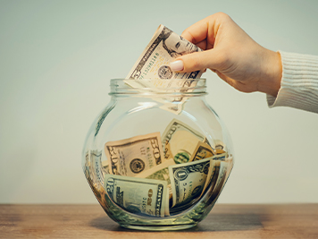 person butting a five dollar bill into a clear jar full of money