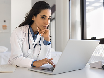 Pensive beautiful young Latin doctor working at laptop in surgery office, touching chin, typing, thinking on consultation in Internet, professional job tasks, medical career, healthcare problems