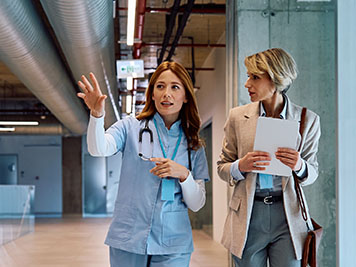 Female nurse and businesswoman talking while walking through hospital hallway. Copy space. 