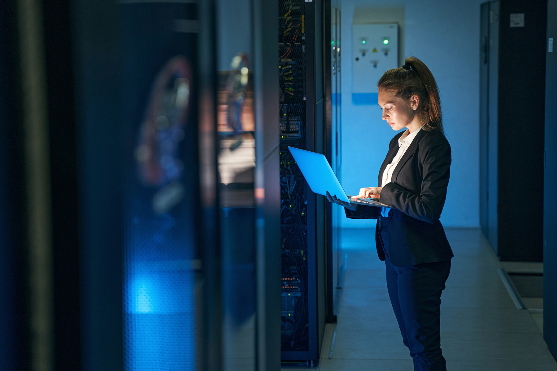 Woman IT specialist in elegant suit working on notebook computer in data center next to server racks