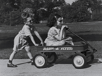 A black and white photo of a toddler pushing another toddler in a wagon