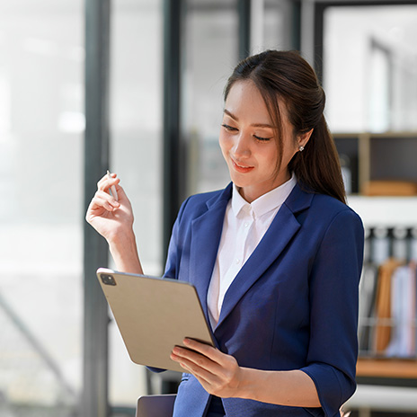 Attractive smiling Asian businesswoman standing holding tablet working and recording work details in office.