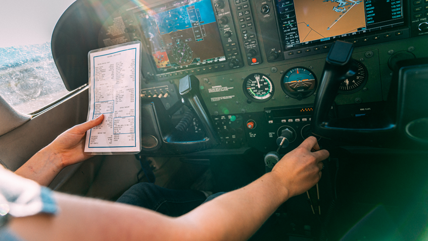 A Zebra named Ed being airlifted via helicopter in a dark mesh sack flying above the trees against blue skies.  